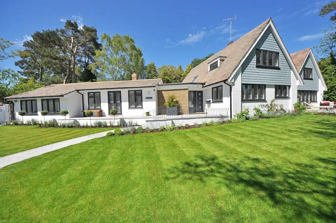 A bright, modern white house with dark-trimmed windows and a green shingled roof, set against a clear blue sky with lush green grass and trees in the foreground.
