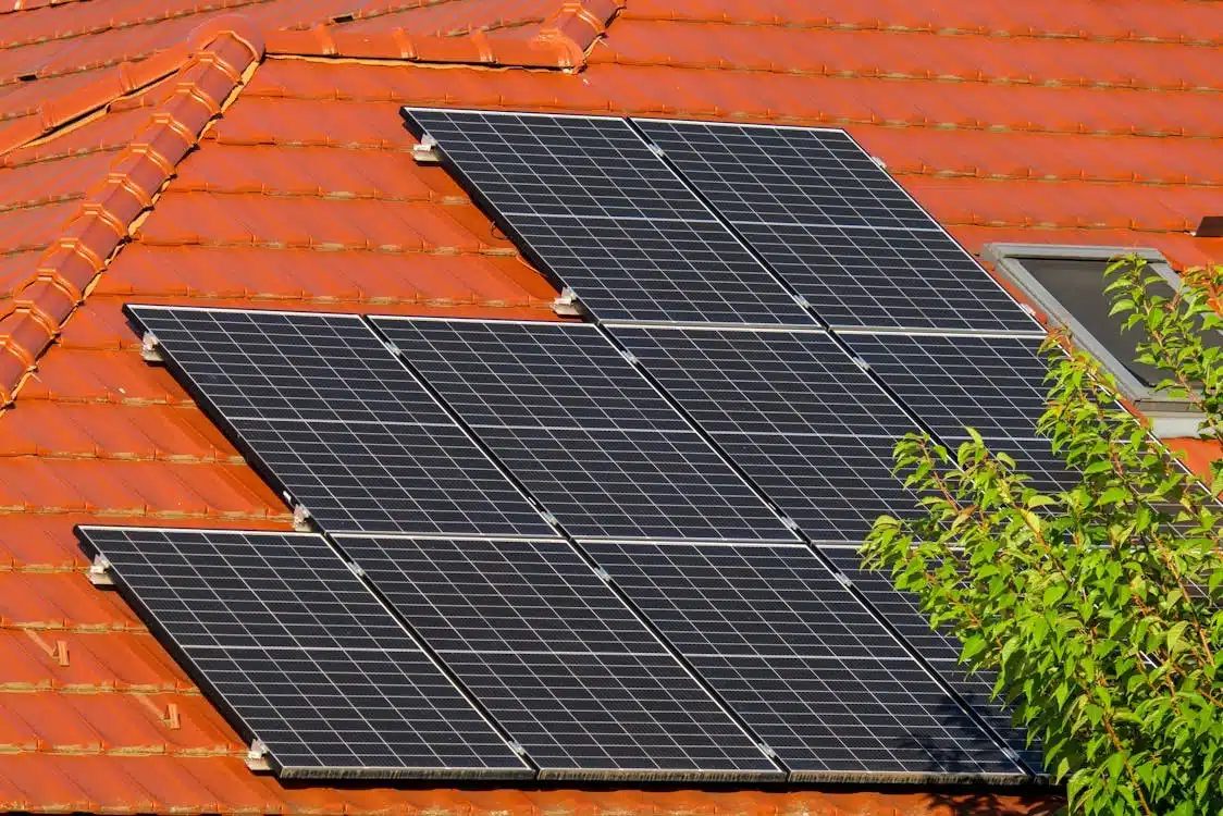 Multiple black solar panels installed on an orange-tiled roof, with a skylight and green foliage visible on the right.