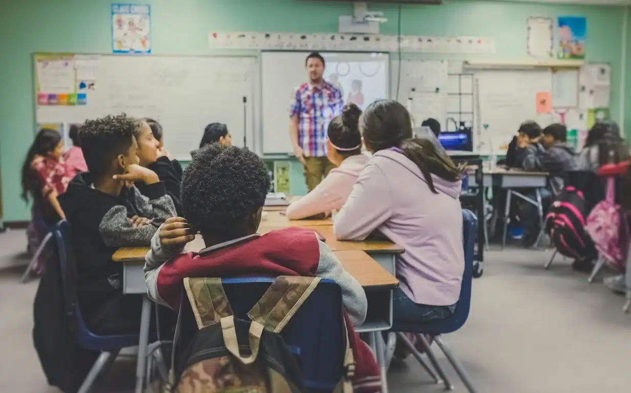 A male teacher stands at the front of a classroom, facing his students who are seated at desks and listening.