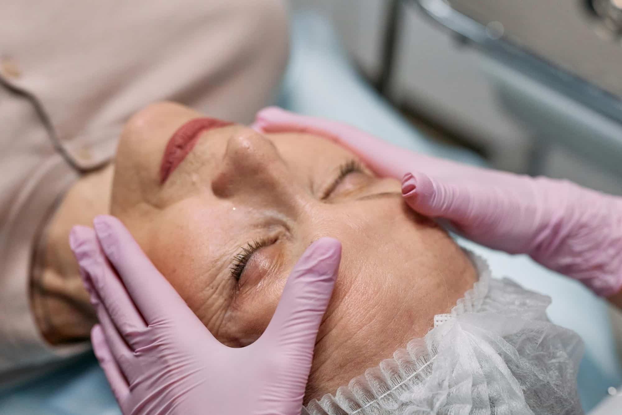 The Future of Non-Invasive Anti-Aging Treatments A person wearing pink gloves gently touches the face of an elderly woman with her eyes closed, indicating a skincare treatment.