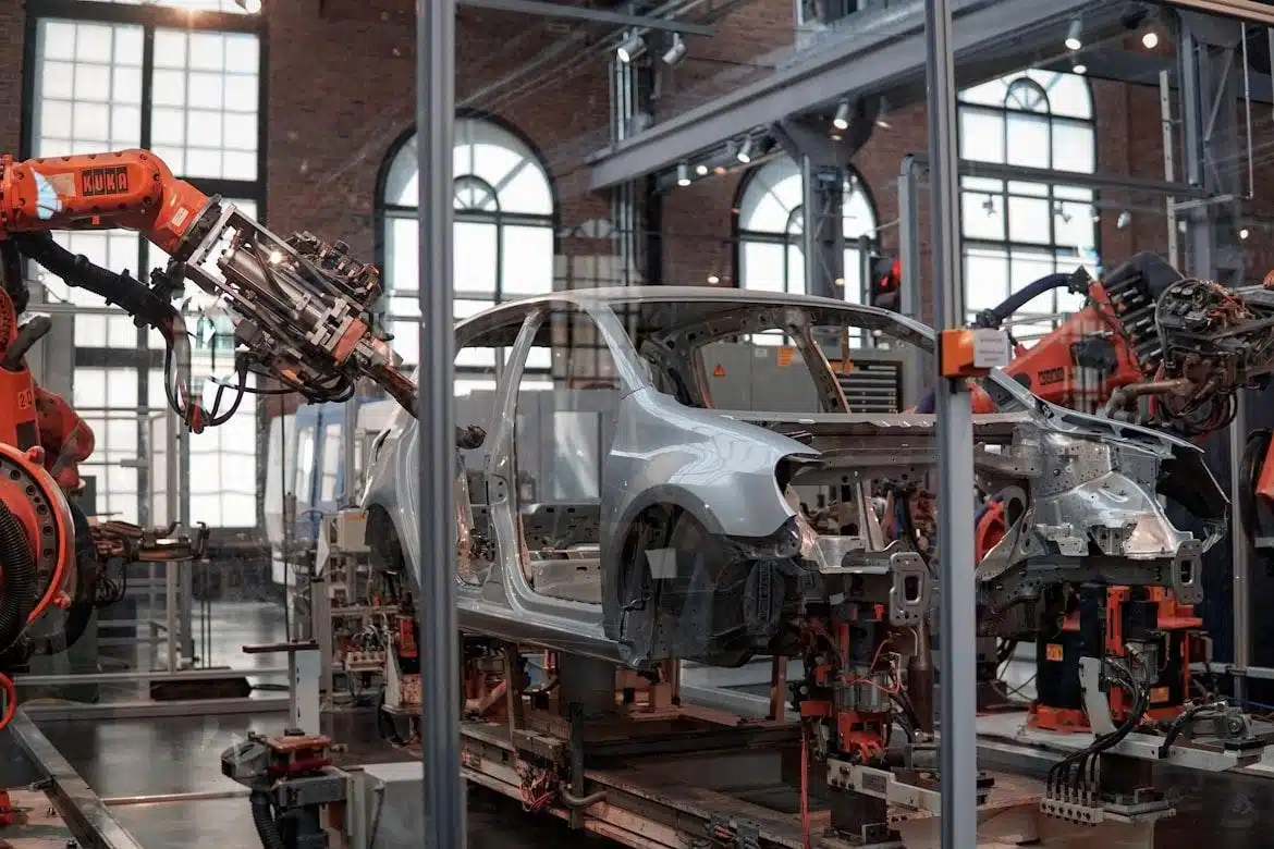An orange KUKA robotic arm works on the frame of a silver car chassis on an assembly line.