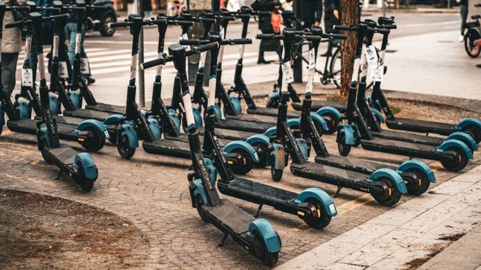 escooter-lineup-urban-transport A row of blue and black electric scooters are parked side-by-side on a cobblestone street.