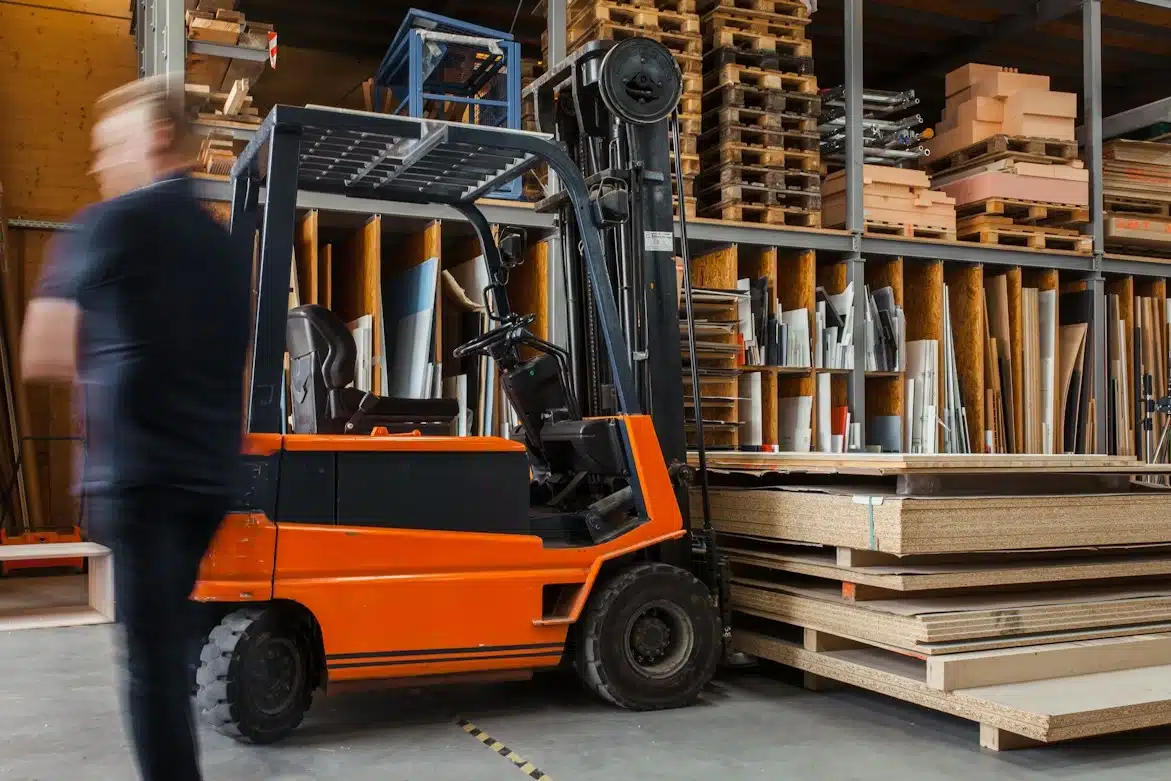 A blurred figure of a person walks past an orange forklift in a warehouse filled with shelves of materials.