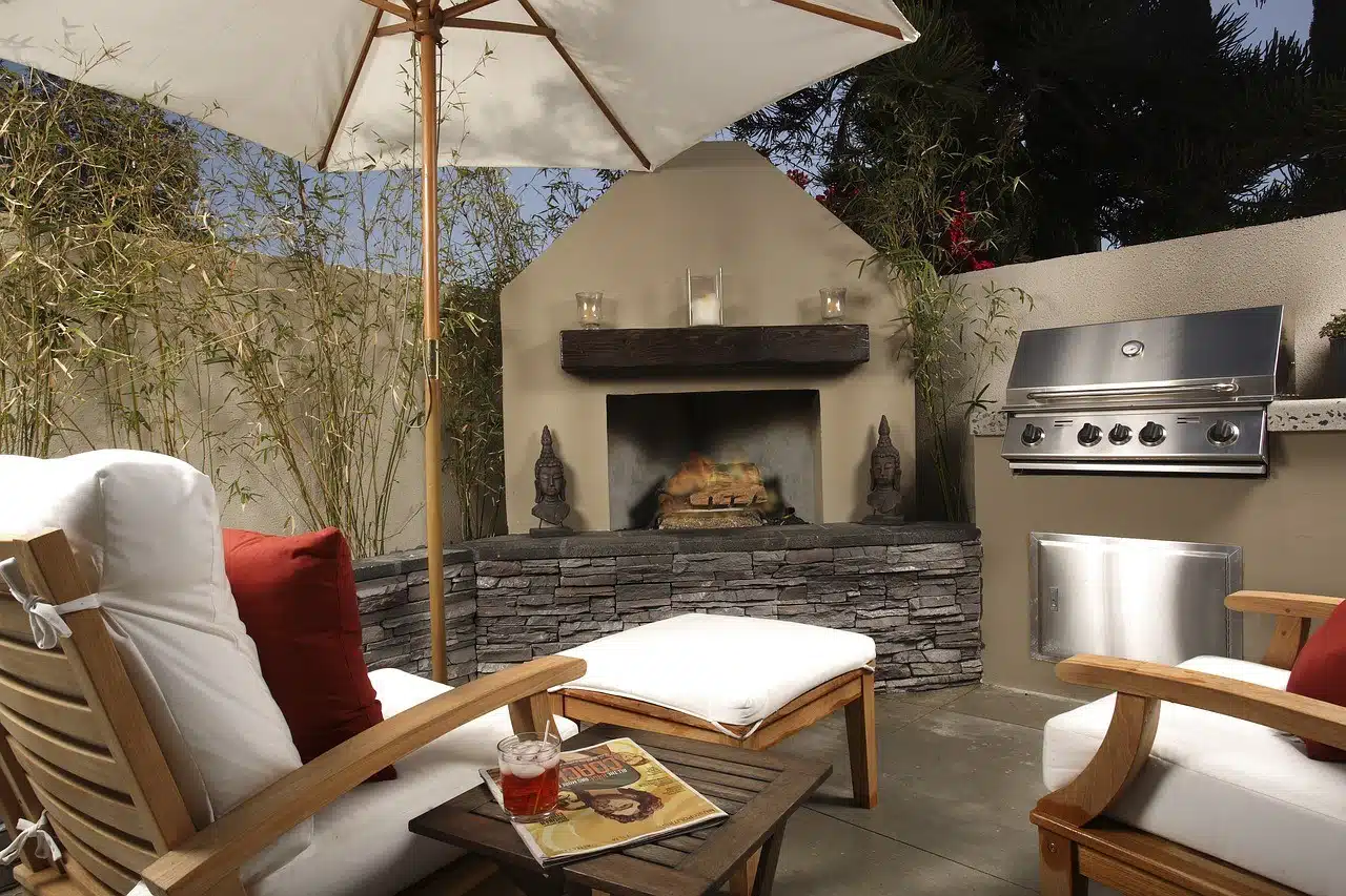 An outdoor patio space features a stainless steel grill and a stone-clad fireplace with a wooden mantle, flanked by two Buddha statues, with comfortable seating and a white umbrella.