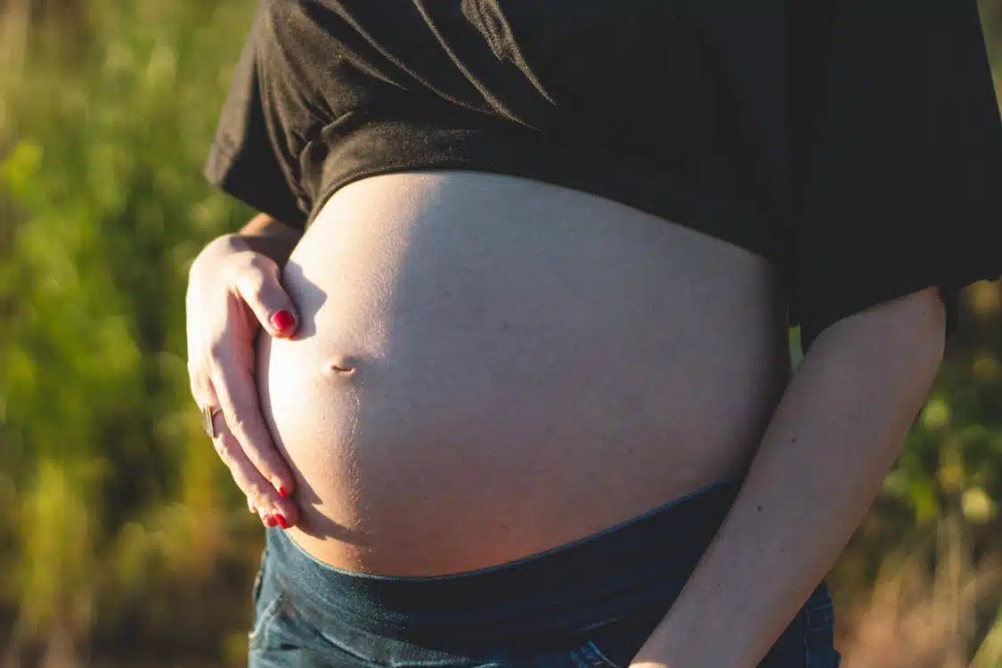 Close-up of a pregnant person's belly, with one hand resting on it, outdoors in dappled sunlight.