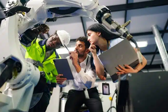 robotics-team-inspecting-machine Three engineers, two men and one woman, are gathered around a large industrial robot. One man, wearing a high-visibility jacket and hard hat, is examining the robot's arm. The other man, in a white shirt, holds a tablet and points towards the robot. The woman, also in professional attire, holds a laptop and appears to be listening intently.