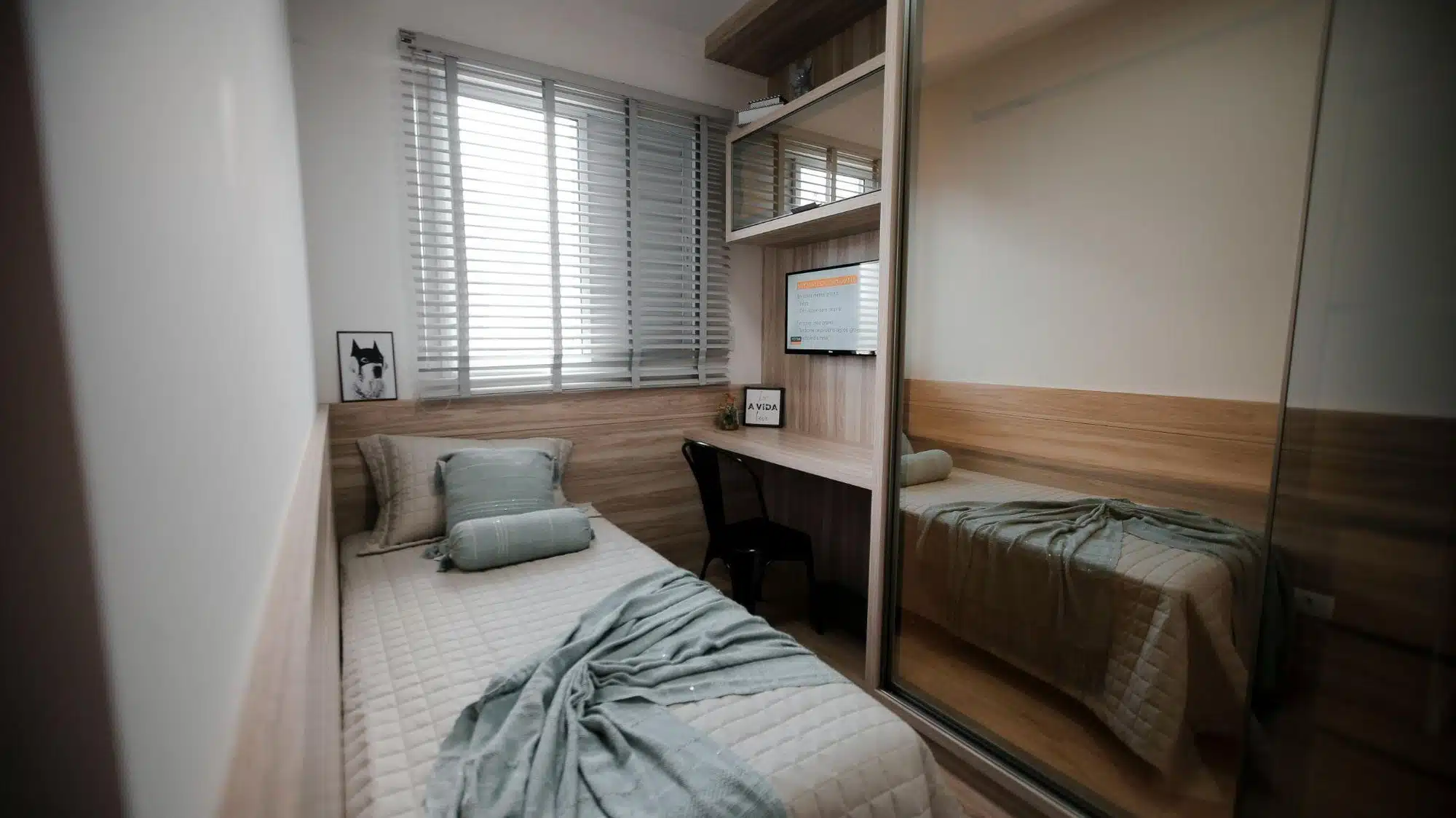 A compact bedroom with a single bed, a desk built into the wall, and a mirrored closet, illuminated by natural light from a window with blinds.