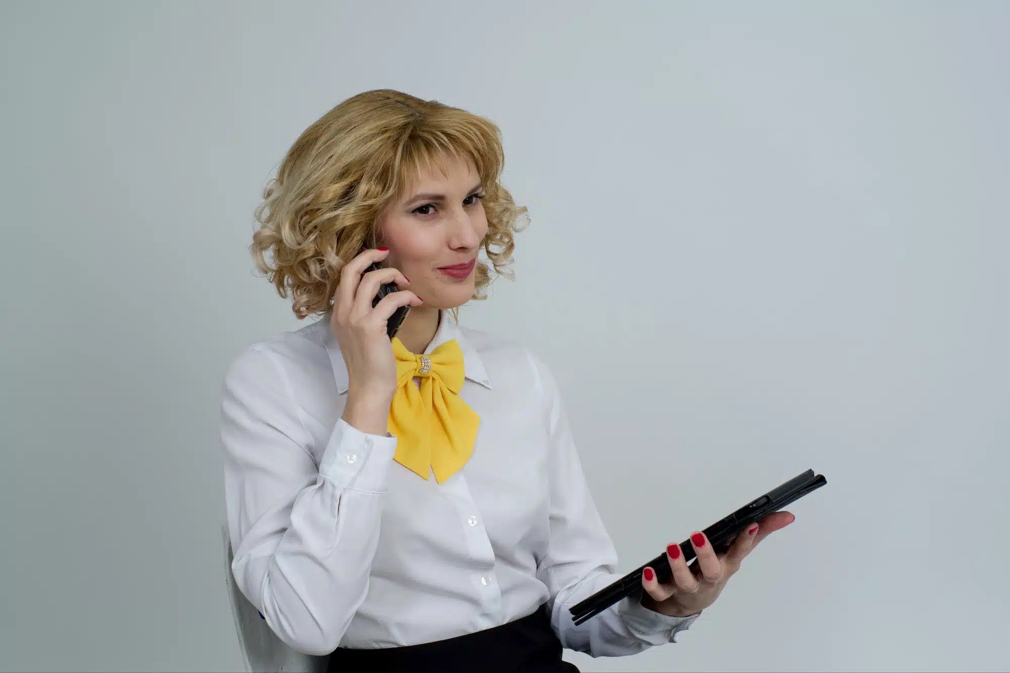 A woman with blonde, curly hair wearing a white blouse with a yellow bow tie is talking on a cell phone while holding a tablet in her other hand.