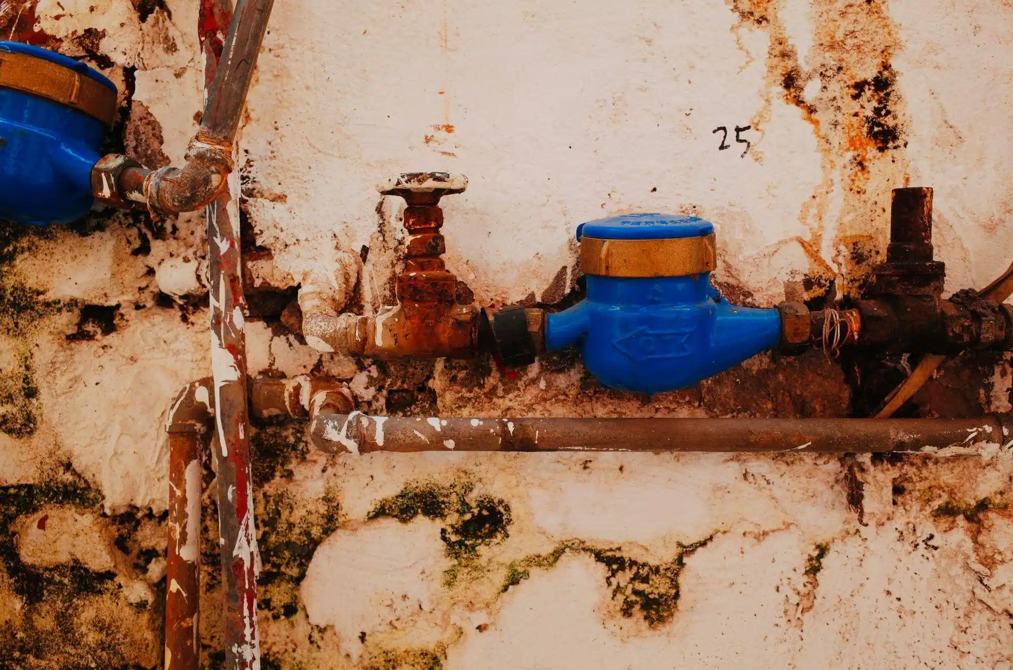 A close-up of a corroded water meter and pipes attached to a dirty wall with a number 25 written in black marker.