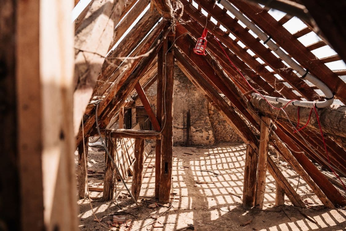 Top 10 Attic Restoration Companies After Wildlife Damage The interior of a damaged attic shows exposed wooden beams and rafters, with sunlight filtering through gaps and casting grid-like shadows on the dusty floor.