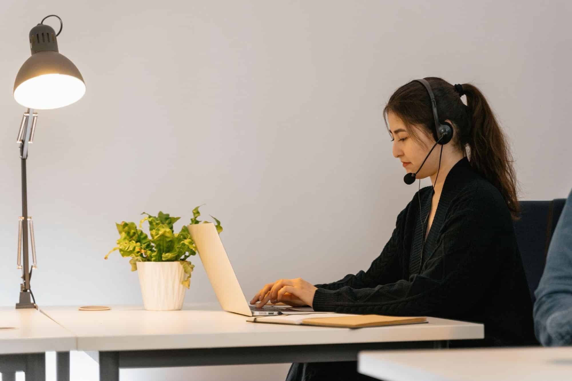 A young woman wearing a headset with a microphone, typing on a laptop at a desk with a lamp and potted plant.