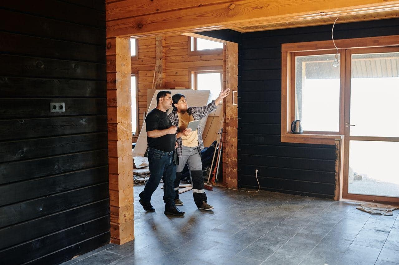 A man and a woman stand in a room undergoing renovations, with the woman pointing towards the wall as she holds a tablet.