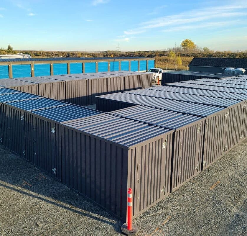 A field of dark grey storage containers with ribbed sides are arranged in a labyrinth-like pattern, with rows of blue-doored storage units visible in the background under a clear blue sky.
