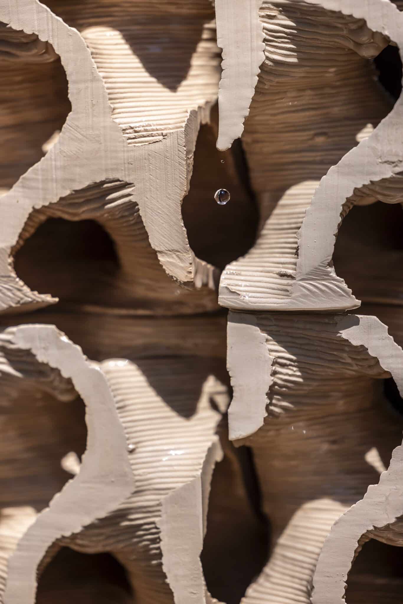 Close-up of a textured, light-brown ceramic sculpture with organic, curved openings. A single water droplet is suspended in mid-air within one of the openings, with smaller droplets visible on the surfaces.