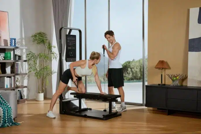 home-gym-workout-couple A man and a woman are working out together in a modern living room. The woman is on a bench, doing a bicep curl with resistance bands attached to a weight machine. The man stands behind her, holding the resistance band and smiling.