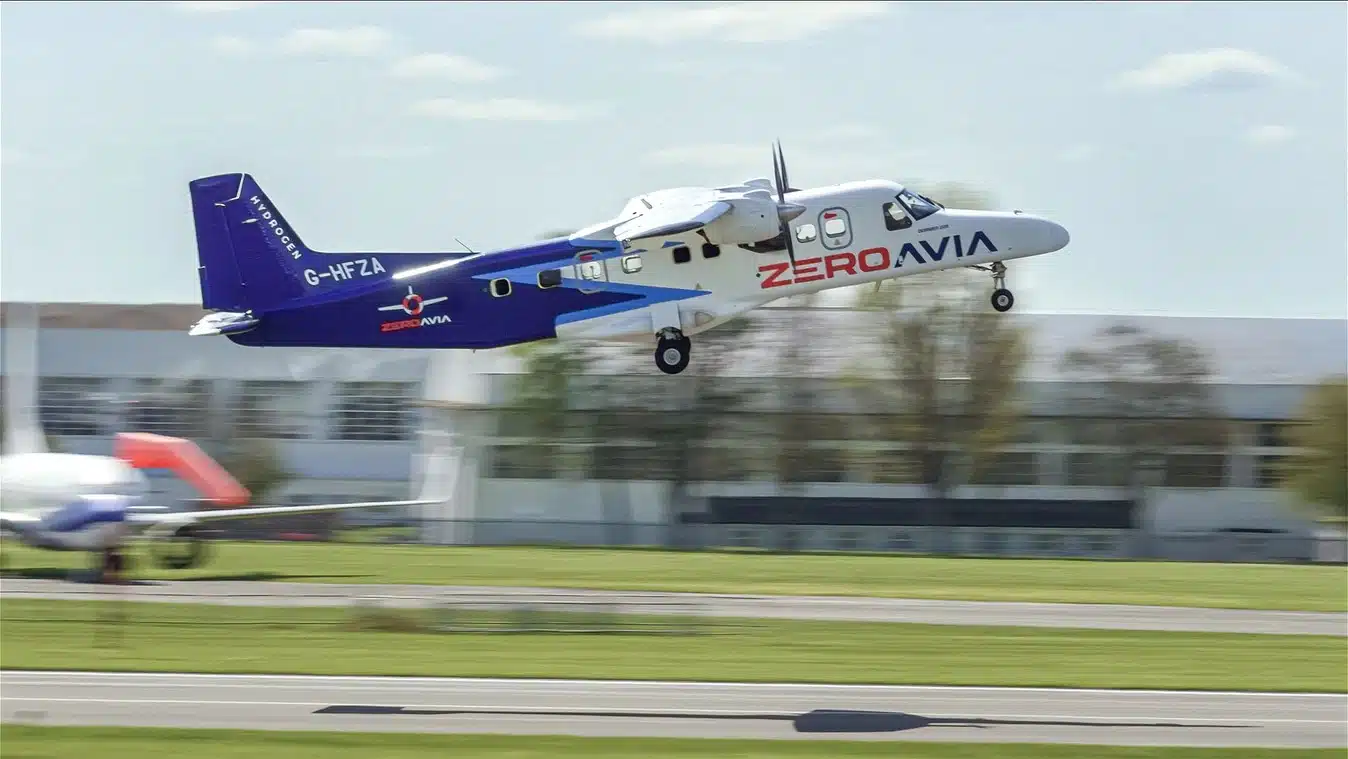 A twin-engine turboprop aircraft with the text "ZEROAVIA" and "HYDROGEN" visible, is captured in mid-air during takeoff. The aircraft has registration "G-HFZA" and is painted in blue and white with red accents. Another smaller aircraft is blurred in the foreground.