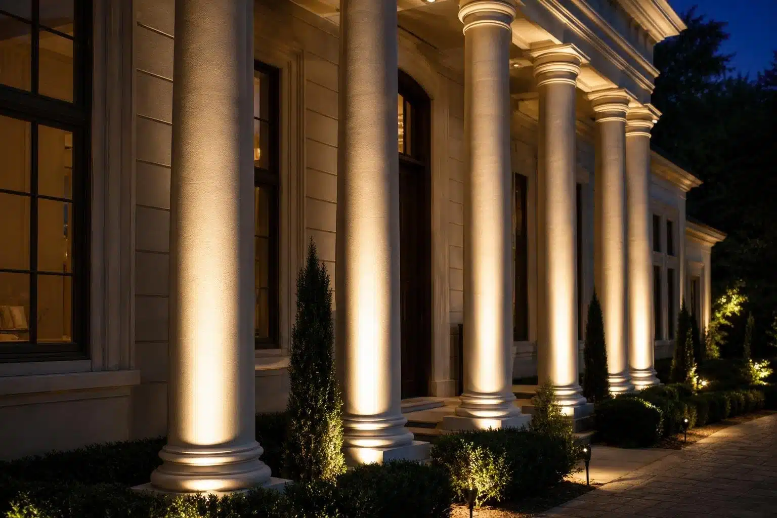 A row of tall, illuminated classical columns on the facade of a building at night. Small evergreen trees and manicured bushes are visible in front of the columns, with pathway lighting adding to the ambiance.