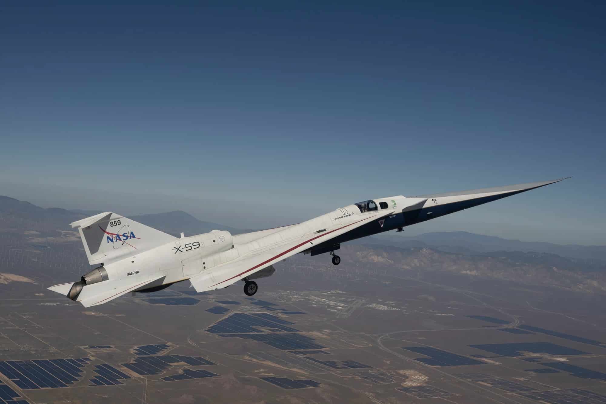 A NASA X-59 aircraft in flight over a desert landscape with solar farms and mountains in the background.