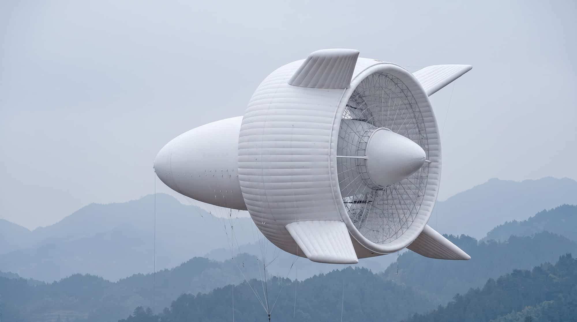 A white rigid inflatable airship with horizontal ribbing, a large rear ducted fan with lattice structure, four stabilizing fins, and a pointed nose cone flying over misty mountains.