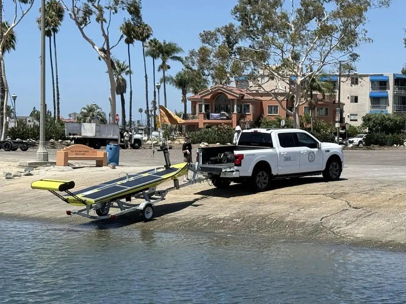 A white SharkLab truck is parked next to a trailer with a yellow and black solar-powered boat on a beach, with palm trees and buildings in the background.