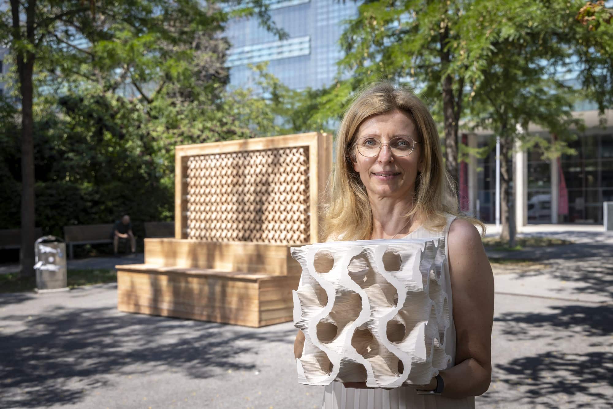 A woman with blonde hair and glasses holds a white, intricate 3D-printed model in her hands. She is standing outdoors, with a wooden bench structure and a building in the background. The model features a complex, organic lattice structure with multiple openings.