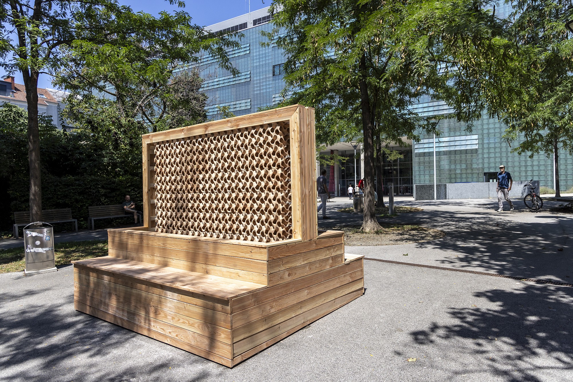 A modern wooden structure with tiered seating and a patterned wooden screen, set in an urban park with a glass-fronted building in the background.