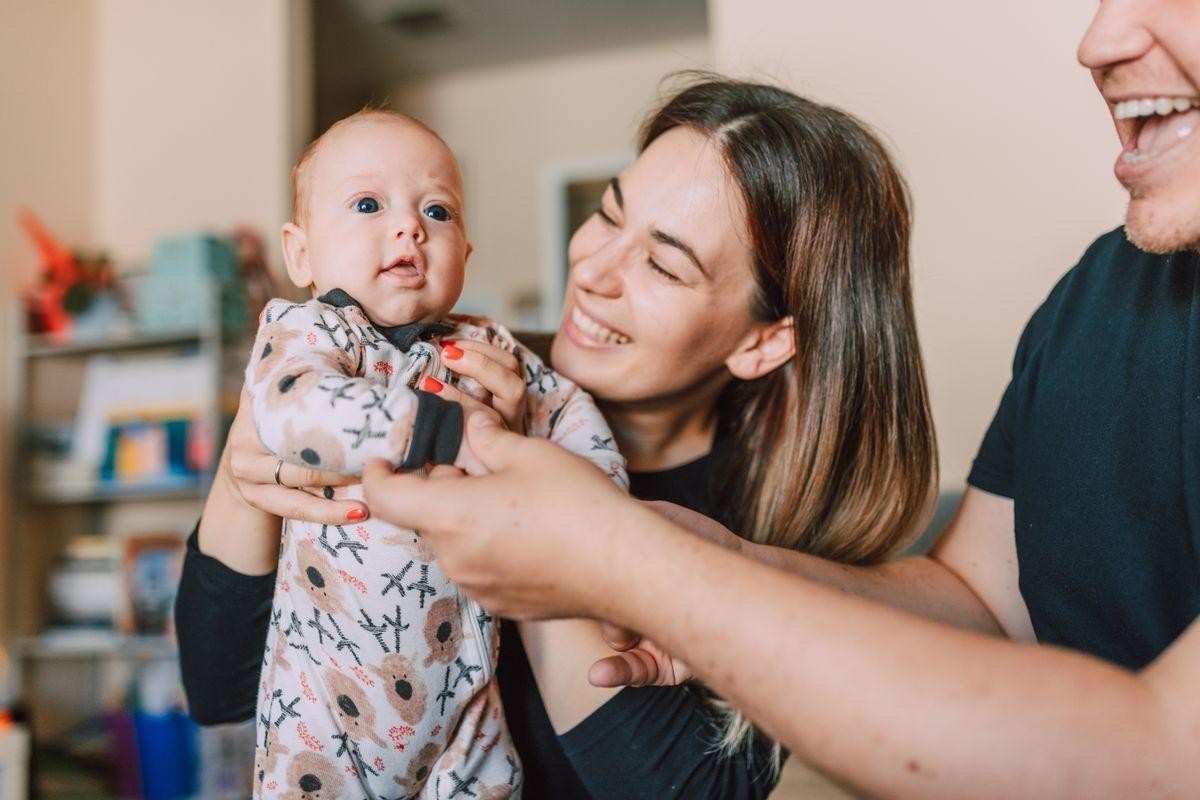 A smiling mother holds her baby while a father, whose mouth is open in a laugh, looks at the baby.