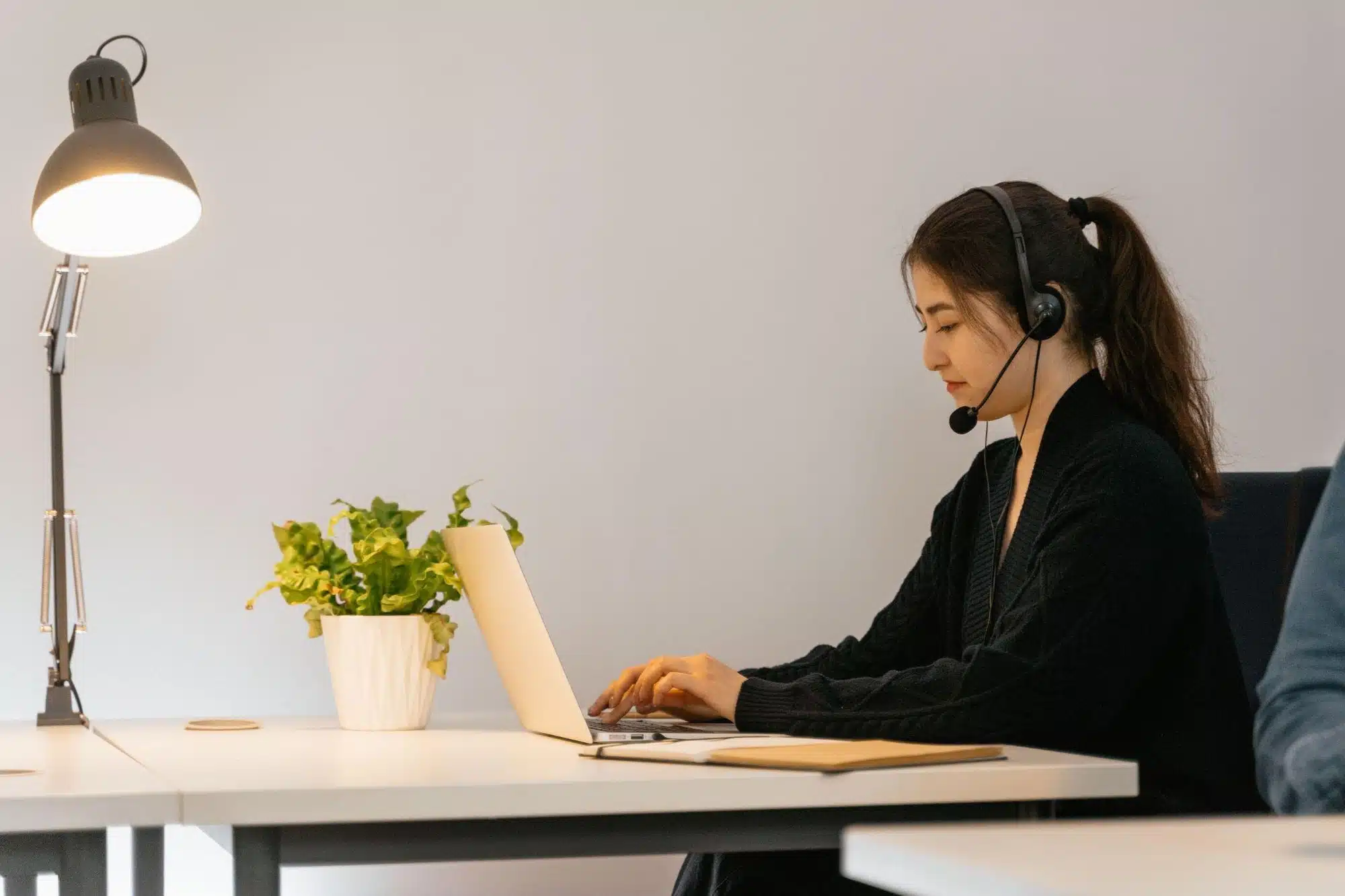 A woman wearing a headset with a microphone types on a laptop at a desk.