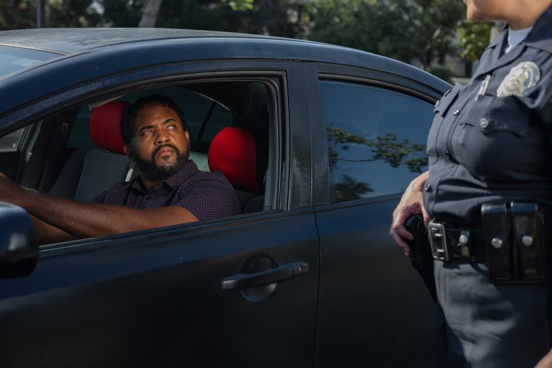 A Black man with a beard drives a car, looking toward a police officer standing outside the car door.