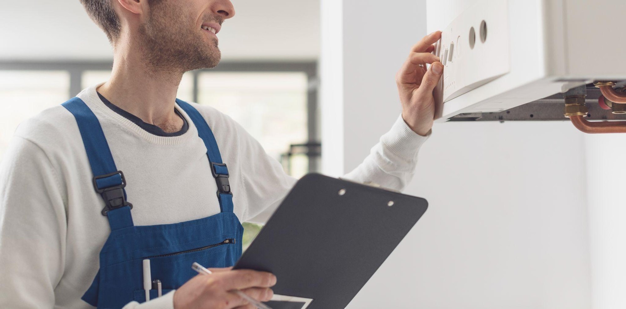 A man in blue overalls and a white sweater is holding a clipboard and pen, adjusting controls on a wall-mounted boiler.