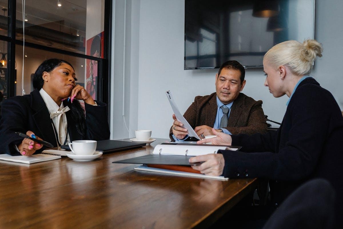 Three people, two women and a man, are seated at a wooden conference table, looking at documents during a meeting.