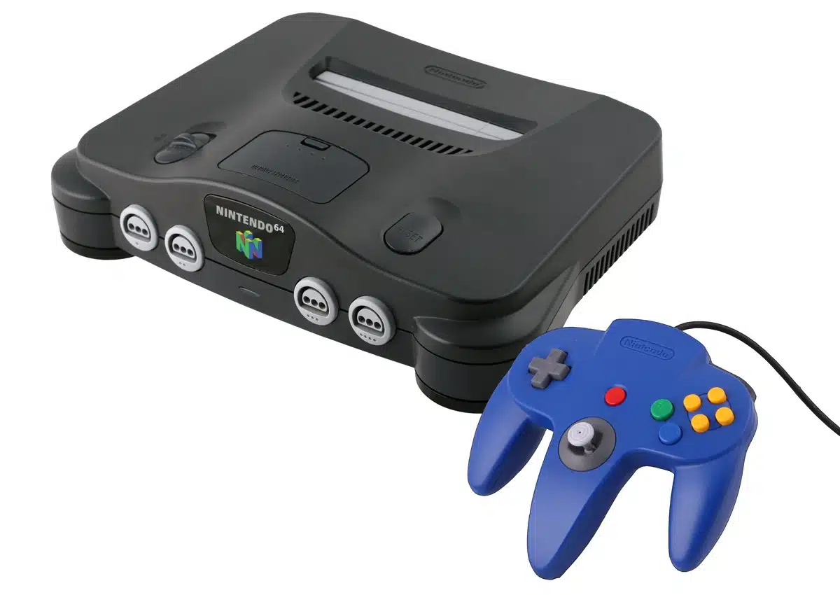 A dark gray Nintendo 64 console is shown from a top-down perspective, with a blue Nintendo 64 controller in the foreground, placed to the right of the console. The console has the Nintendo 64 logo and text on its front panel, and several controller ports. The controller has a distinct three-pronged design with a directional pad, analog stick, and buttons.