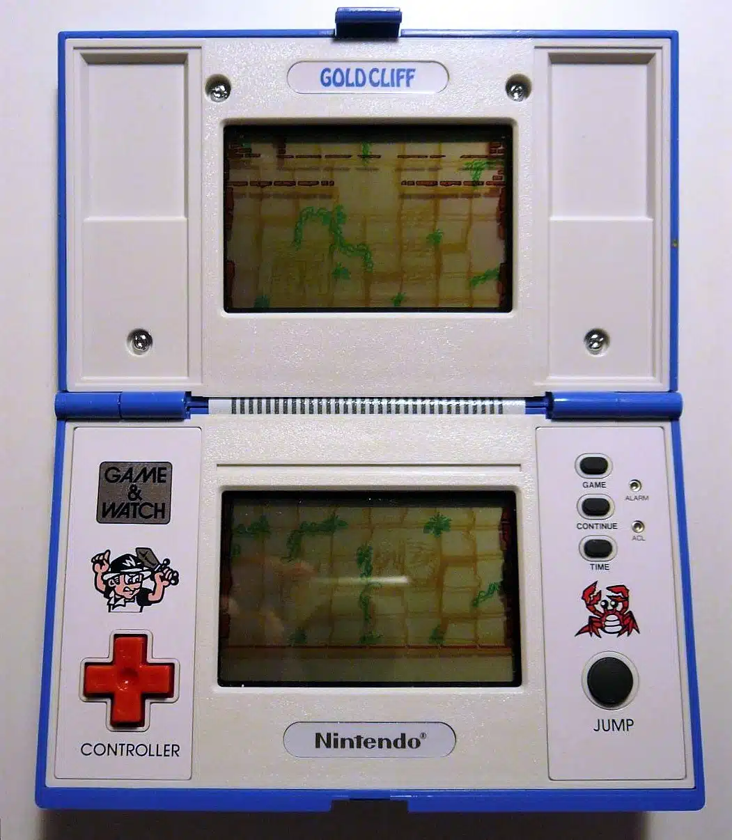 A blue and white Nintendo Game & Watch Gold Cliff handheld electronic game, open and displaying gameplay on its dual screens. The top screen shows a scene with a figure in a helmet and a hammer, and the bottom screen shows a crab-like character. The device has buttons labeled Game, Alarm, Continue, Time, and Jump, and a red directional cross.