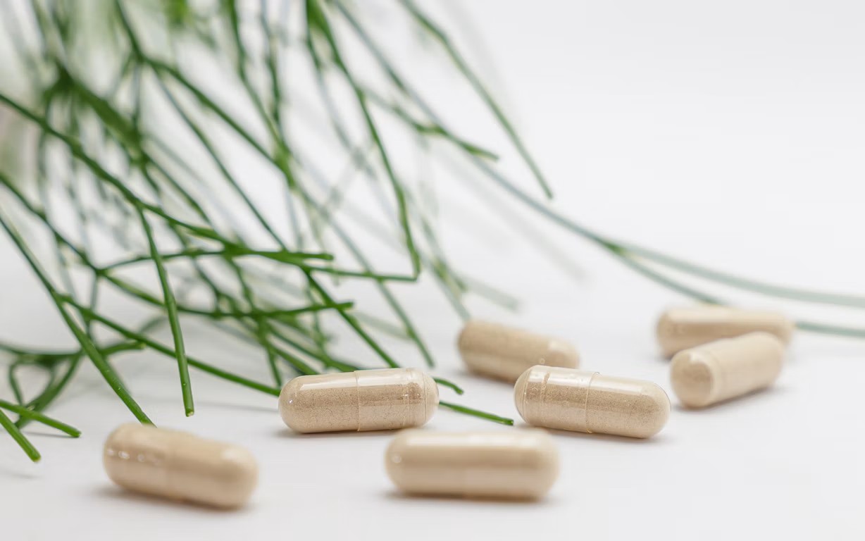 Several beige supplement capsules scattered on a white surface next to green herbal sprigs.