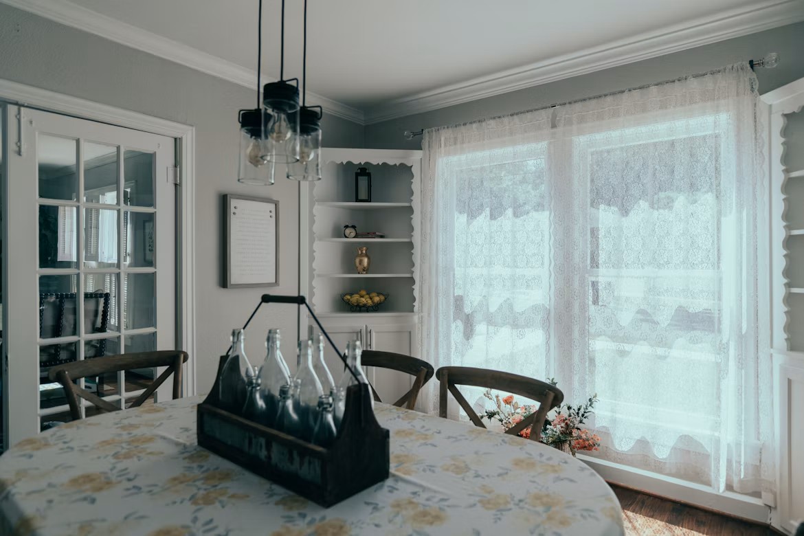 A bright dining room table set with a floral tablecloth and a wooden carrier holding glass bottles near a large window covered in lace curtains.