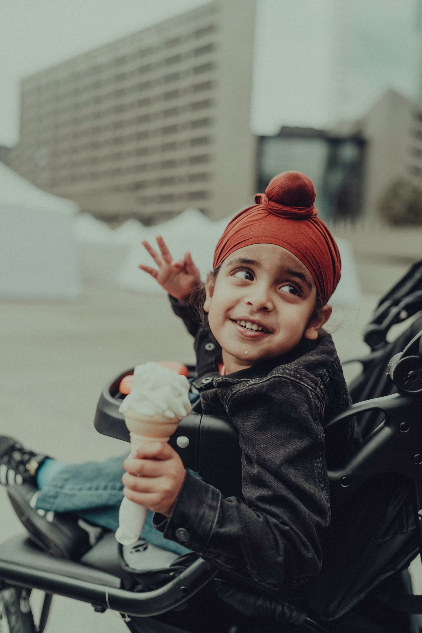 A young child wearing a red patka and holding an ice cream cone while sitting in a stroller outdoors.