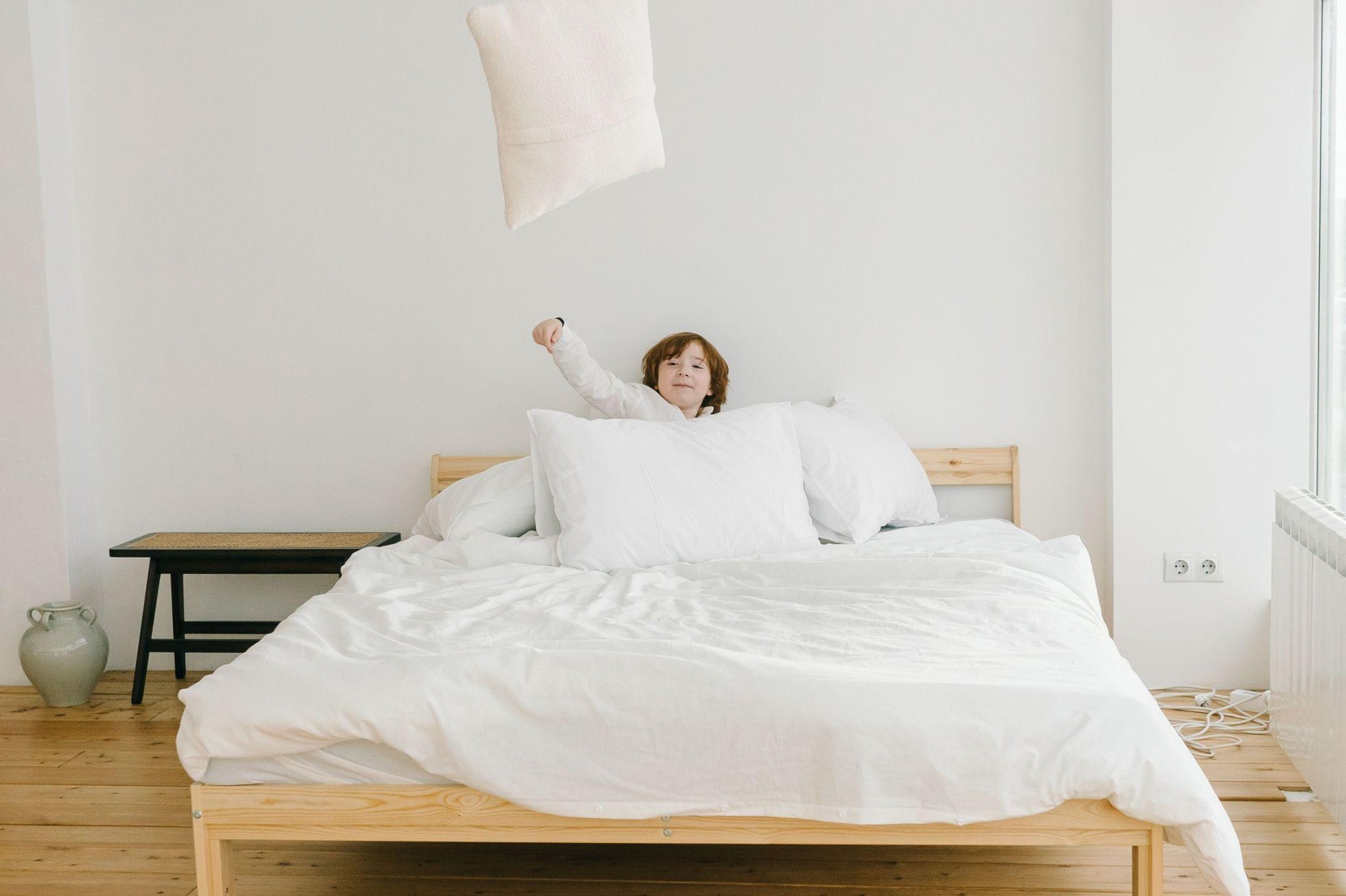 A young child plays on a large wooden bed with white linens, tossing a pillow into the air.