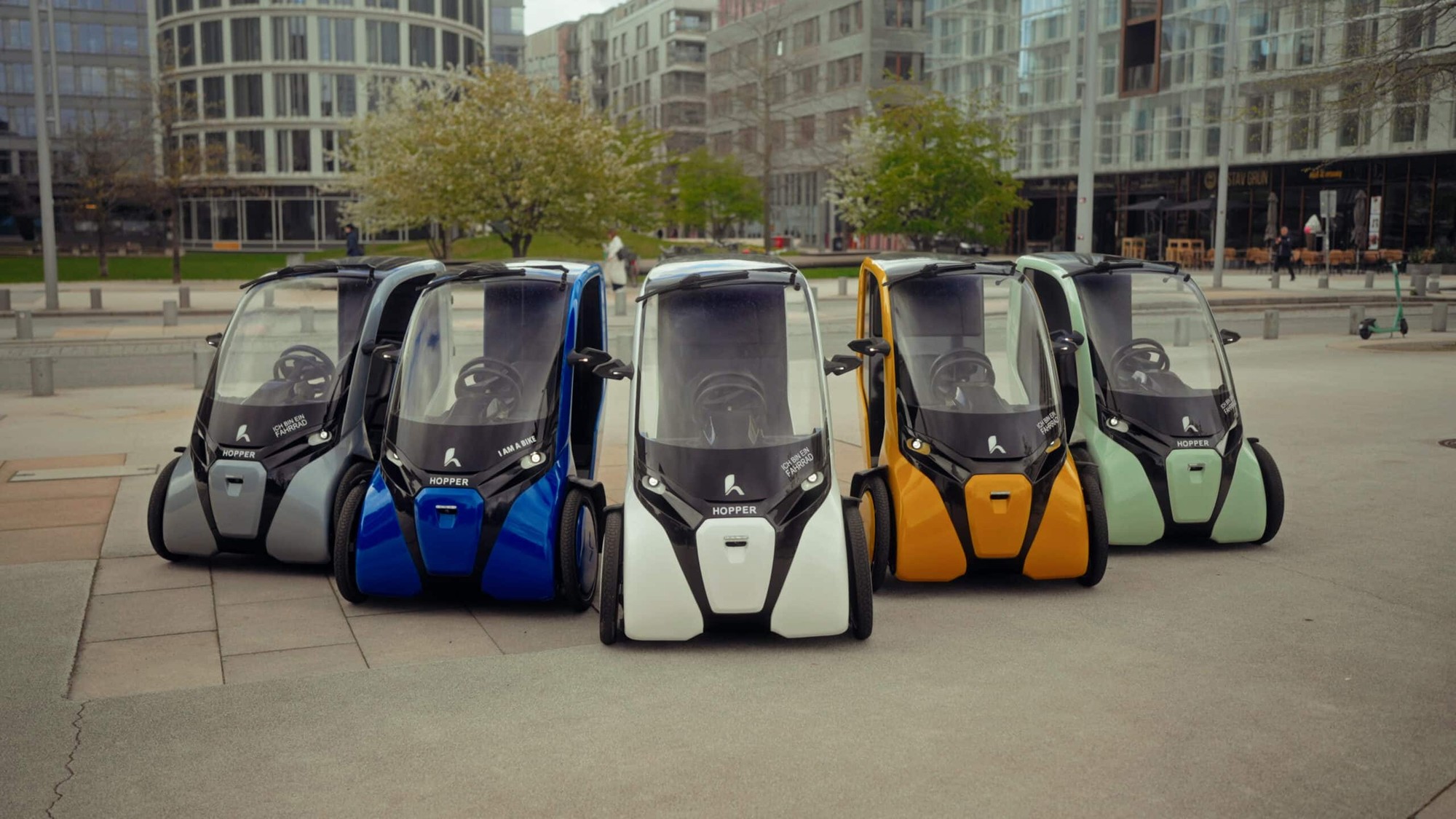 Five colorful Hopper three-wheeled electric vehicles parked in a row on a paved plaza.