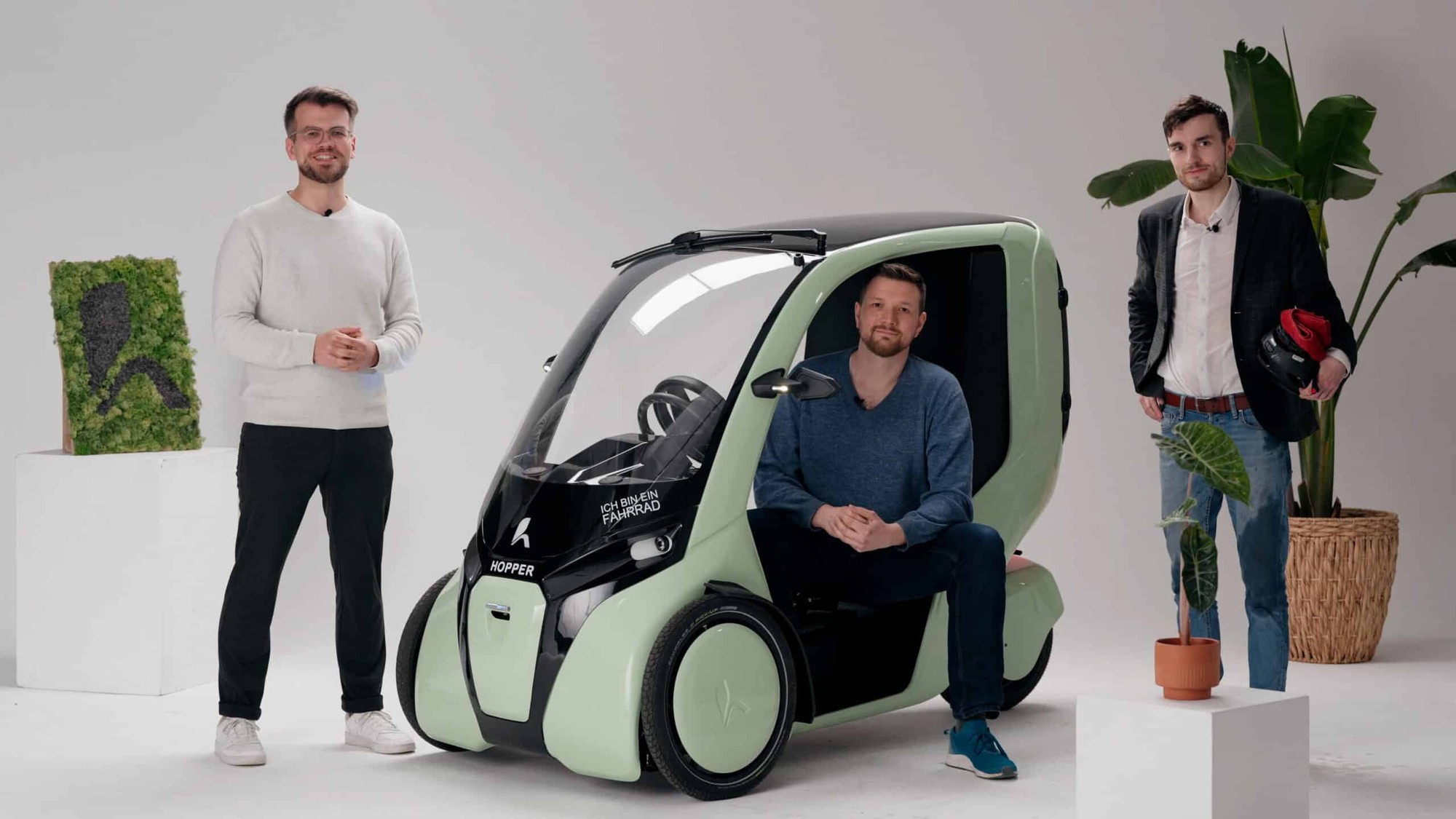 Three men standing beside a light green Hopper printed with the words ICH BIN EIN FAHRRAD.