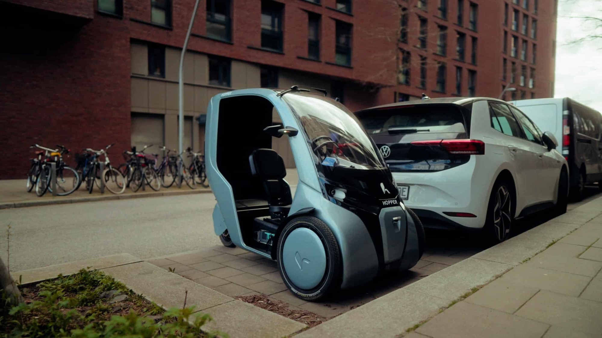 A pale blue Hopper cargo trike parked on a sidewalk next to a white car.