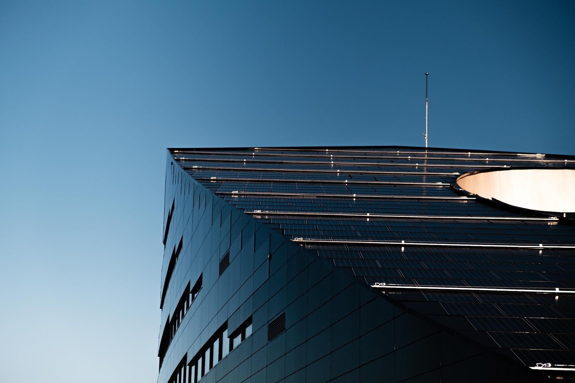 A modern industrial distribution center building featuring solar panels mounted across its angled roof against a clear blue sky.