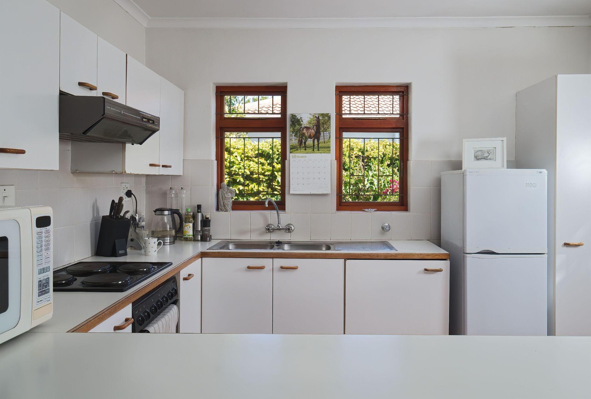 White flat-front cabinets and a white refrigerator in a small kitchen, with a built-in oven and stovetop under a range hood beside a sink set below two wooden-framed windows.