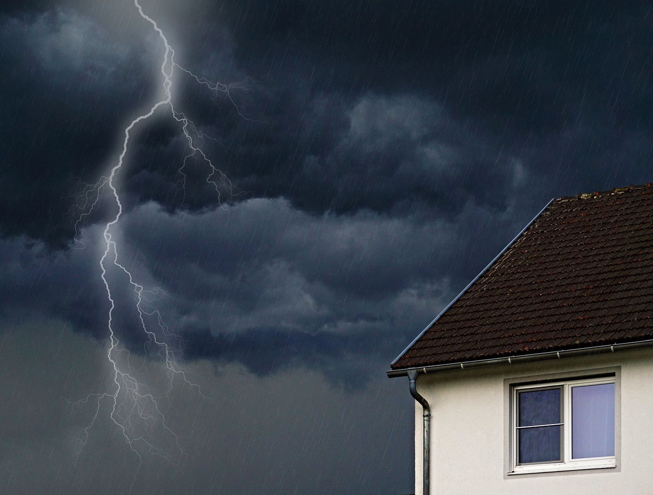 A severe thunderstorm with lightning striking a dark, rainy sky above the corner of a residential home.