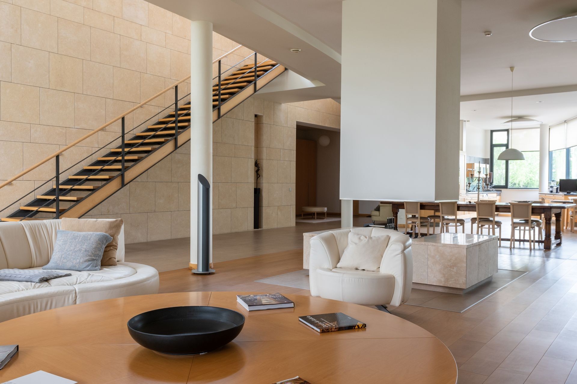 How Digital Conveyancing Is Changing the Cost of Selling a Home in the UK A modern living room with a round wooden coffee table, white leather sofa and armchair, a central white structural column or fireplace partition, a floating staircase on the left, and a dining area in the background.