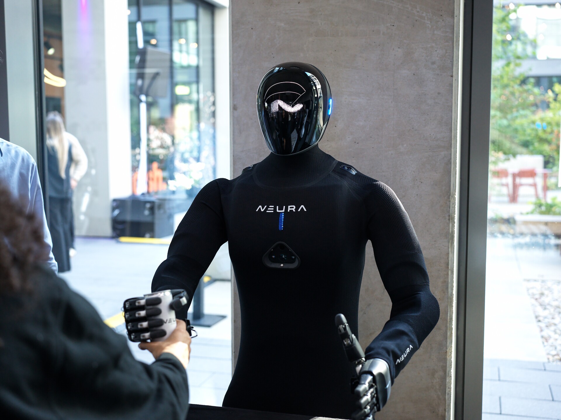 A black humanoid robot by Neura Robotics with a sleek helmet and glowing face design interacting with a person shaking its hand.