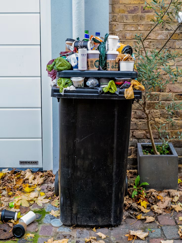 An overflowing black outdoor garbage bin piled high with disposable coffee cups and various refuse on a paved courtyard.