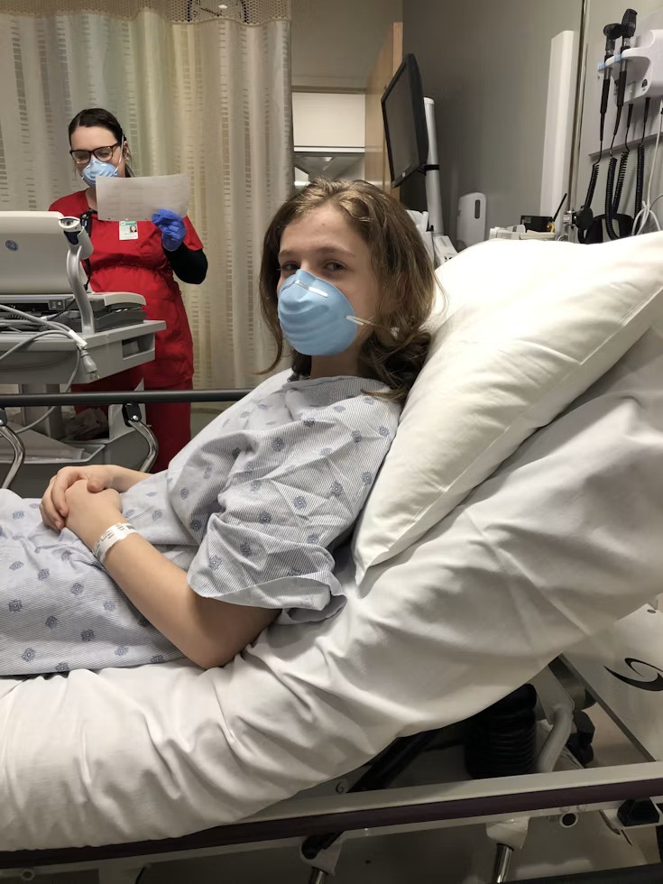 A young patient wearing a face mask sits up in a hospital bed.