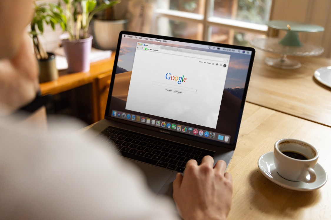 A person using a MacBook Pro with the Google search engine open on screen while sitting at a wooden table with coffee.