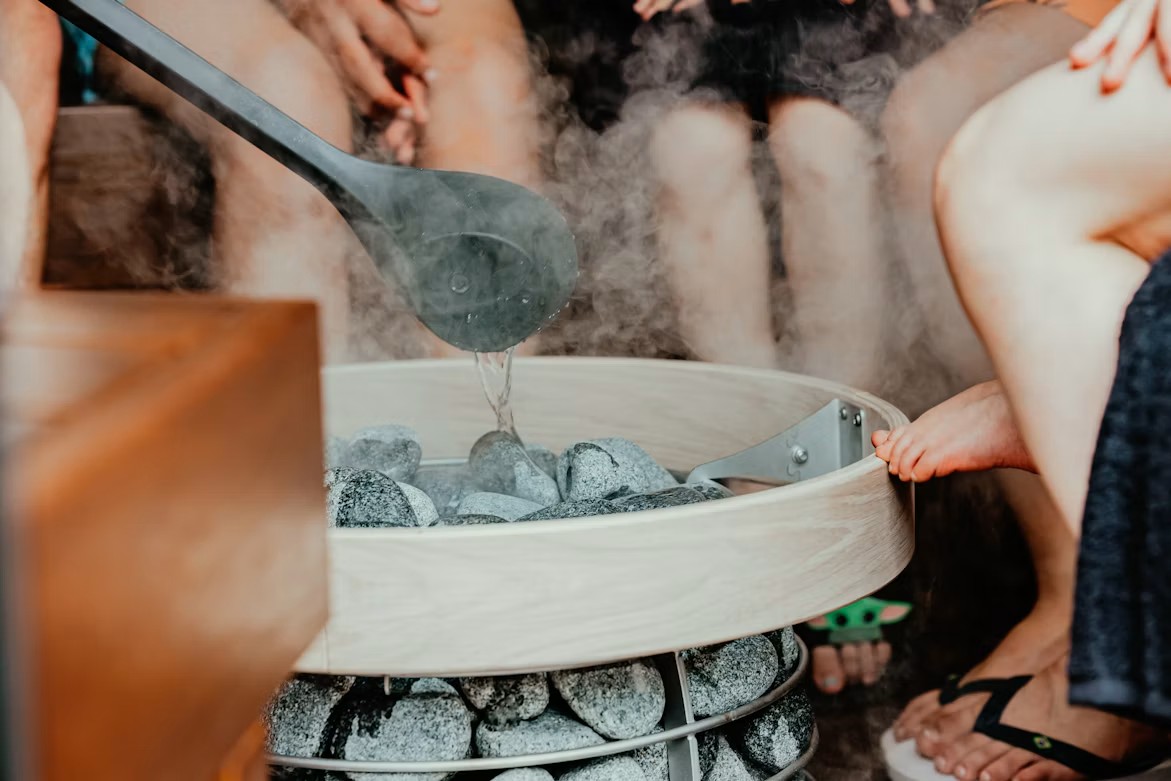A ladle pouring water over hot sauna stones, creating steam in a traditional wooden heater surrounded by people relaxing.
