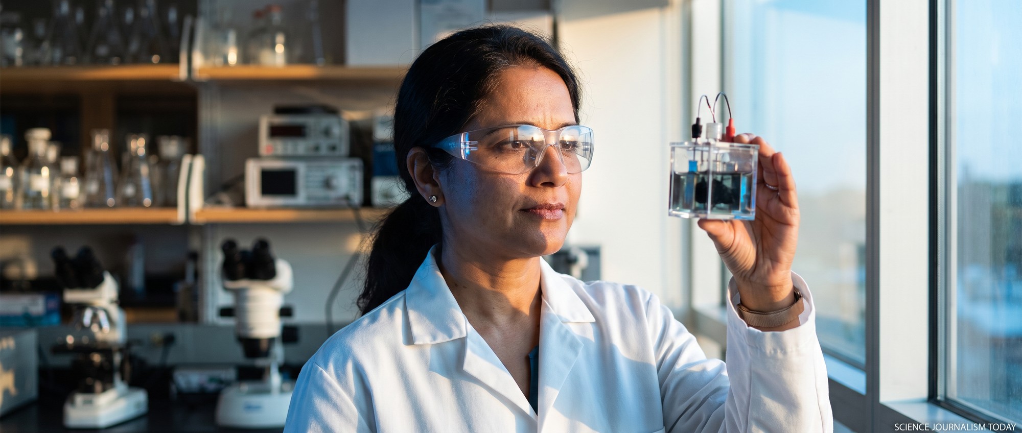 Scientist in a lab coat examining a small bio-electrochemical cell held up to the light